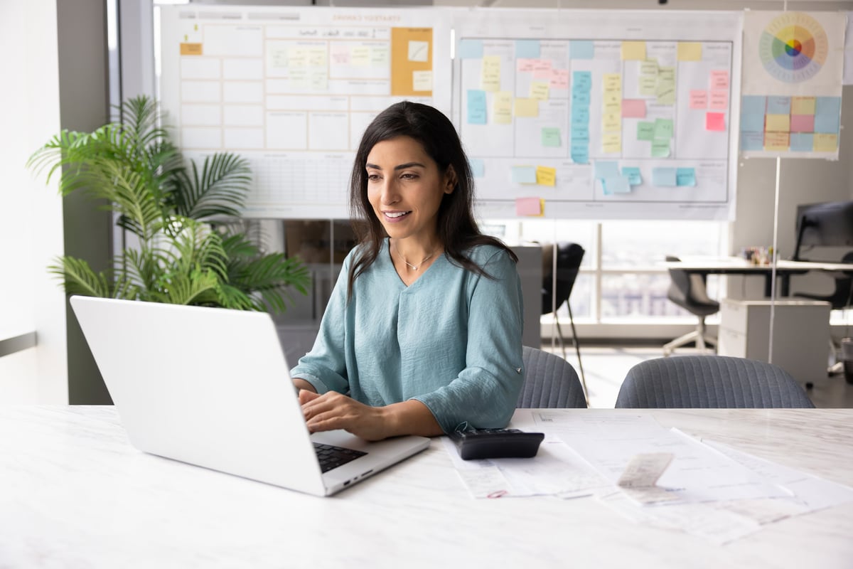 Mujer latina dueña de un pequeño negocio revisando una calculadora, libreta de costos y laptop con Excel abierta, calculando el precio correcto de sus productos con una expresión de confianza